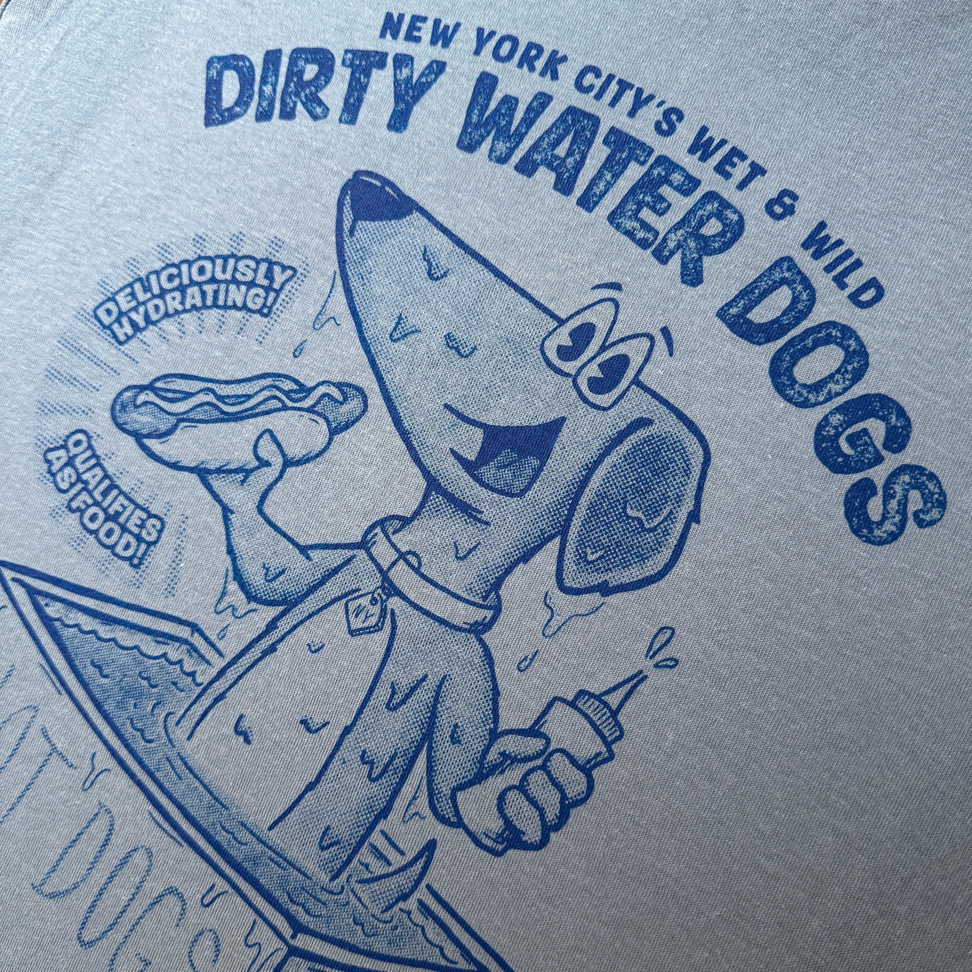 Blue t-shirt with blue graphic and text of a dog holding a bottle labeled 'Dirty Water Dogs' on a wooden surface.