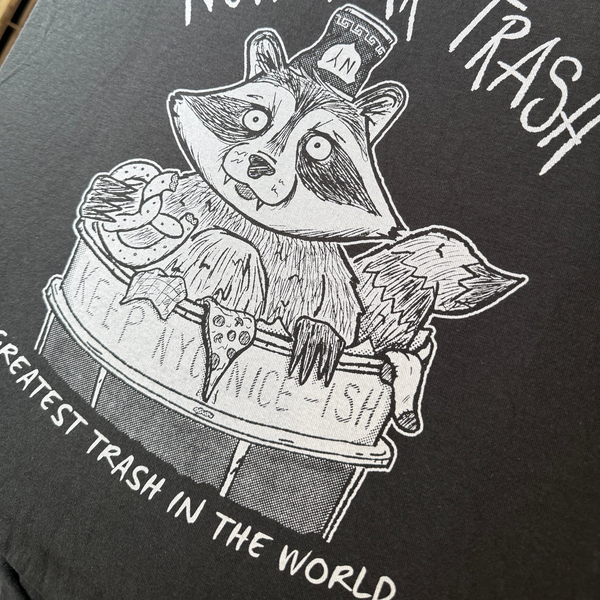 Black and white illustration of a raccoon in a New York City trash can on a dark t-shirt