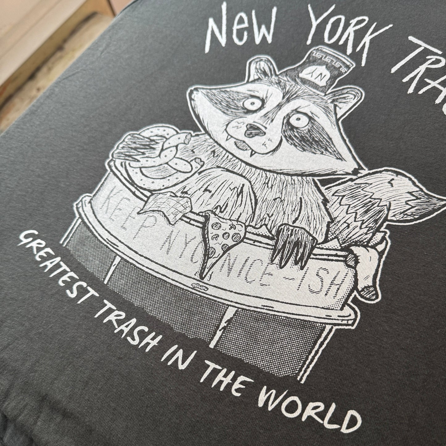 Black and white illustration of a raccoon in a New York City trash can on a dark t-shirt