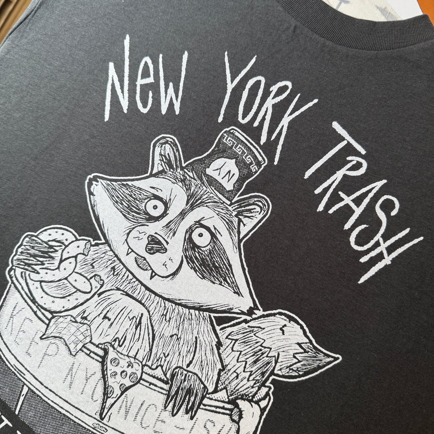 Black and white illustration of a raccoon in a New York City trash can on a dark t-shirt