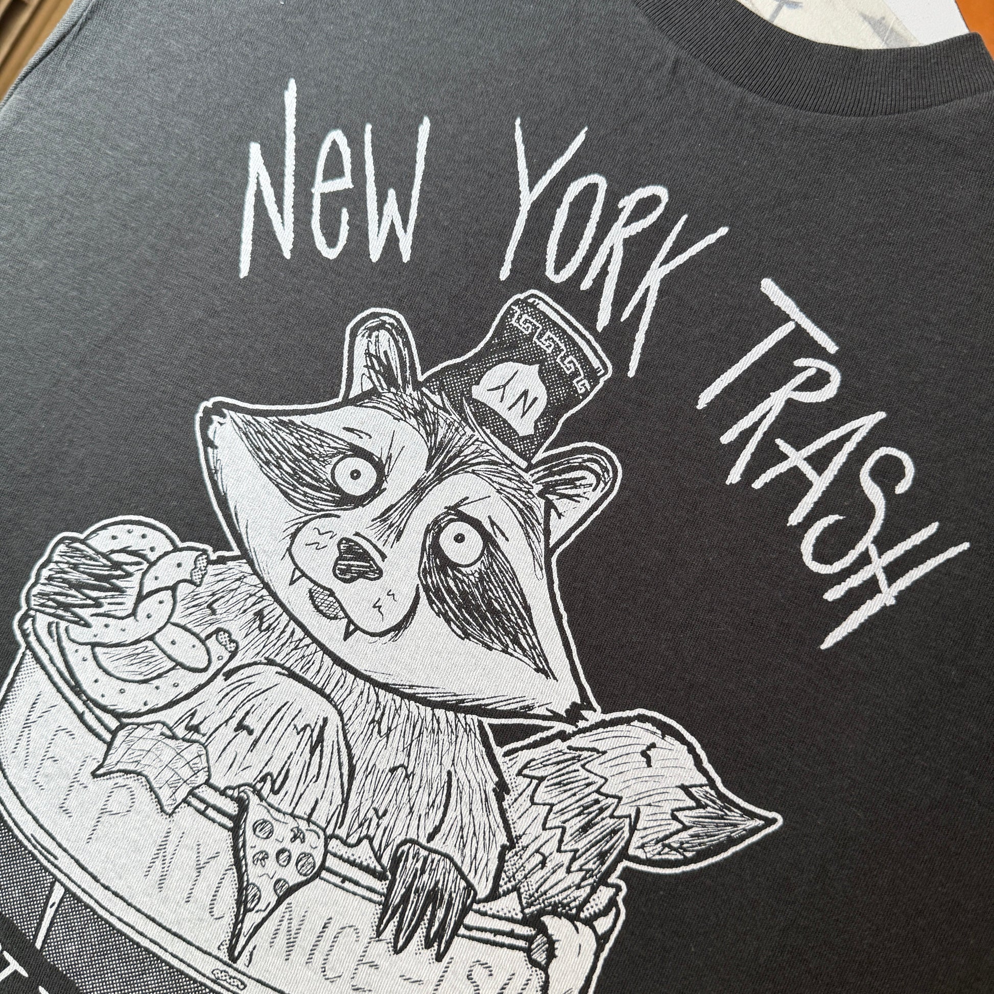 Black and white illustration of a raccoon in a New York City trash can on a dark t-shirt
