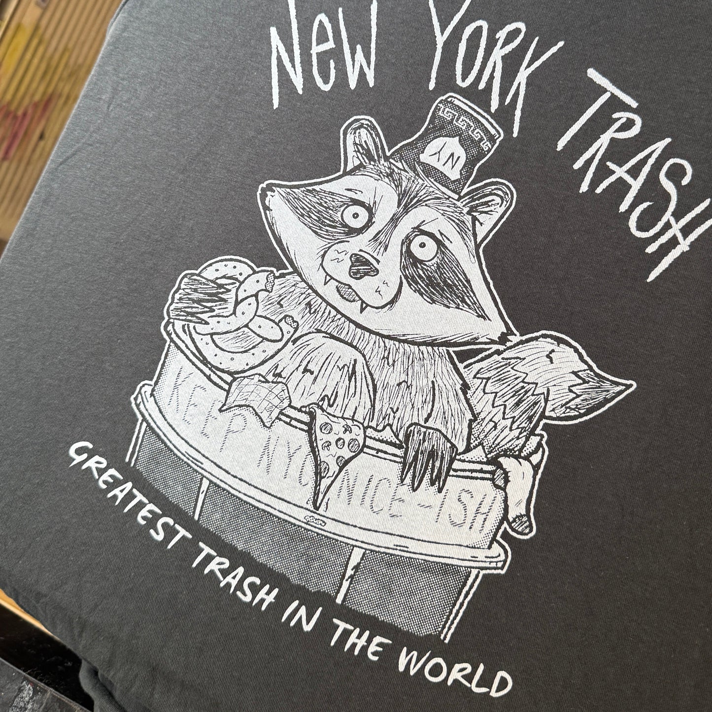Black and white illustration of a raccoon in a New York City trash can on a dark t-shirt