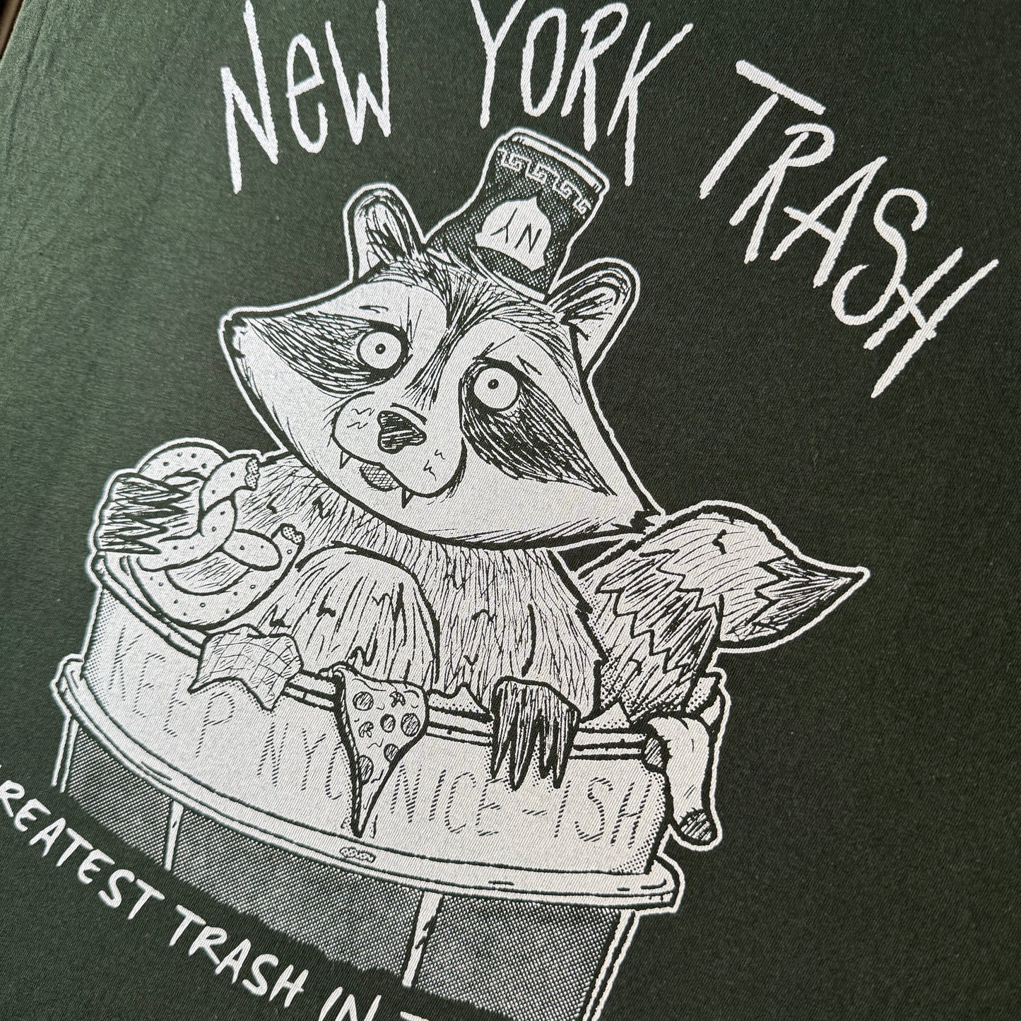 Close-up of a dark green graphic t-shirt featuring a white hand-drawn illustration of a raccoon sitting in a trash can, with the text 'New York Trash' and 'Greatest Trash in the World'.