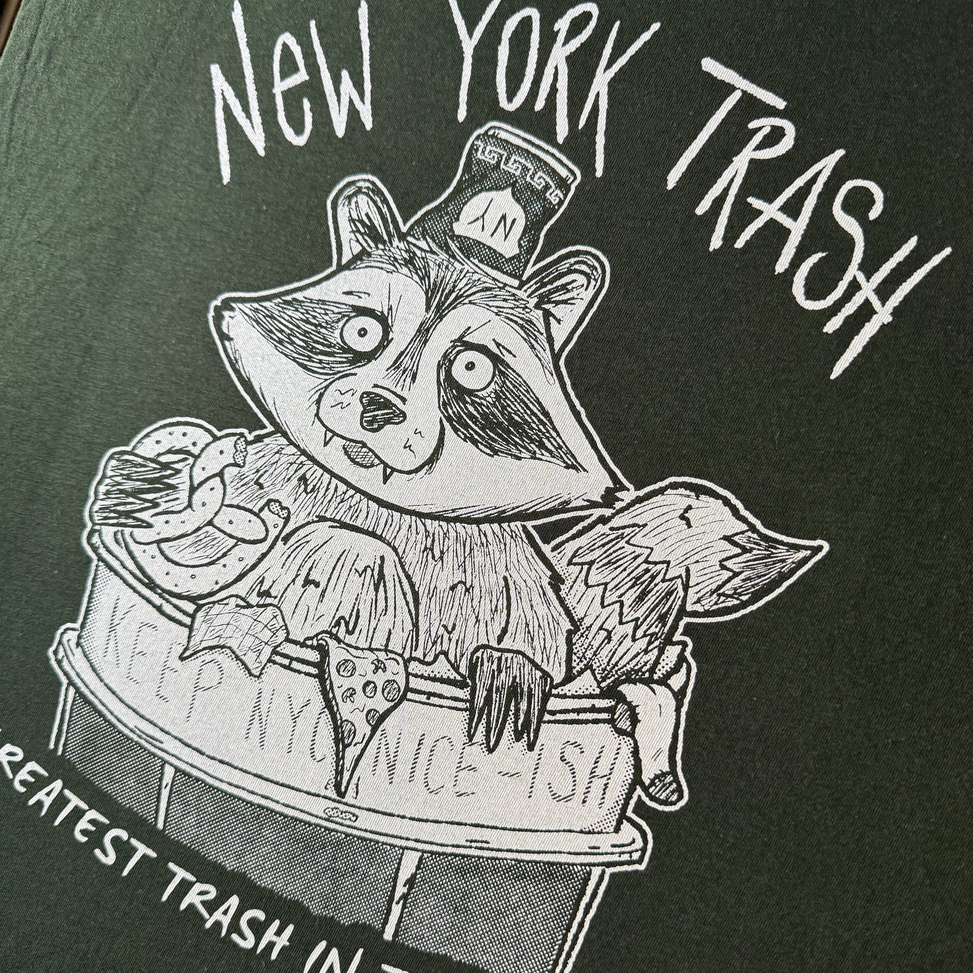 Close-up of a dark green graphic t-shirt featuring a white hand-drawn illustration of a raccoon sitting in a trash can, with the text 'New York Trash' and 'Greatest Trash in the World'.