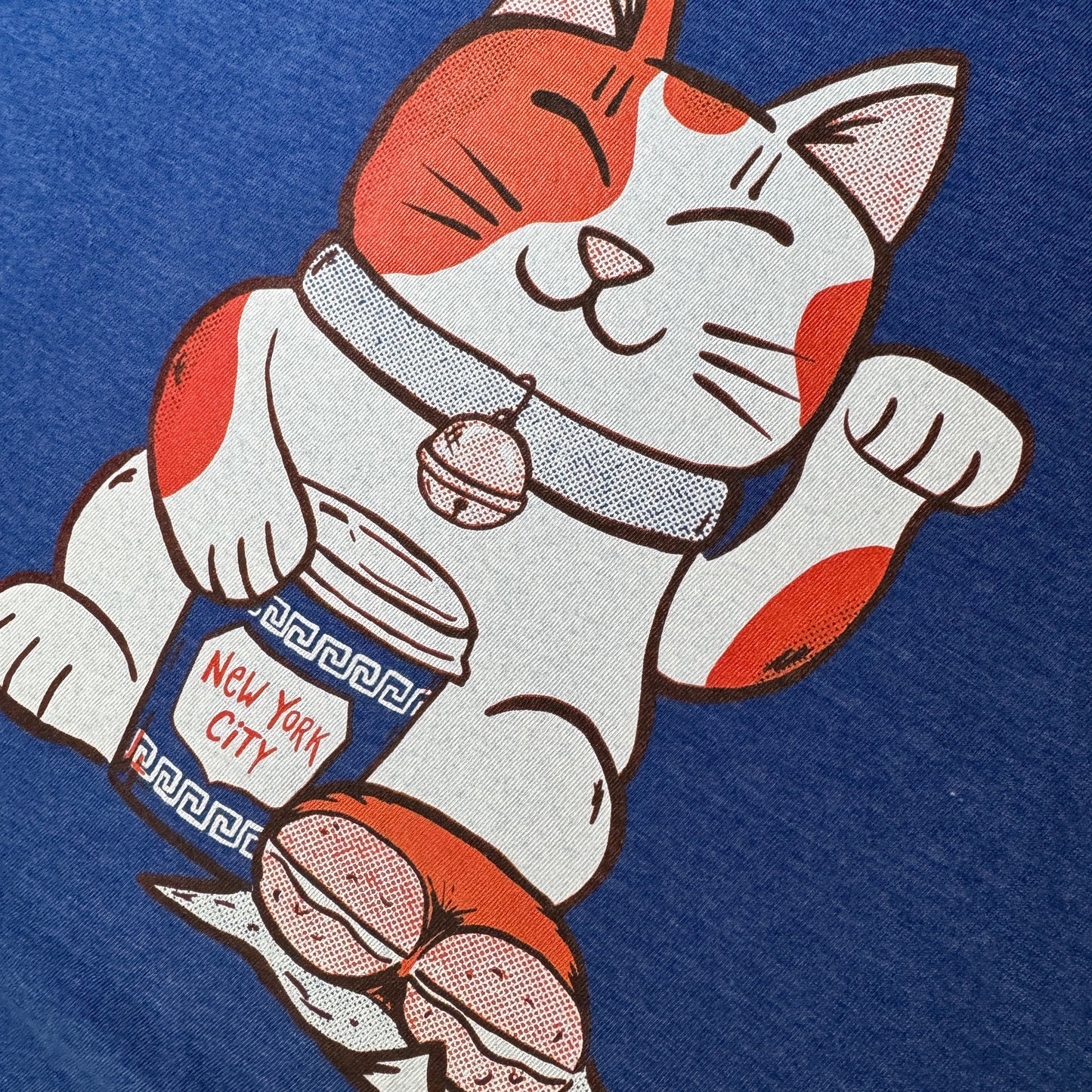 A close-up of a blue t-shirt featuring a printed cartoon maneki-neko cat holding a coffee cup labeled 'New York City' and a bagel.