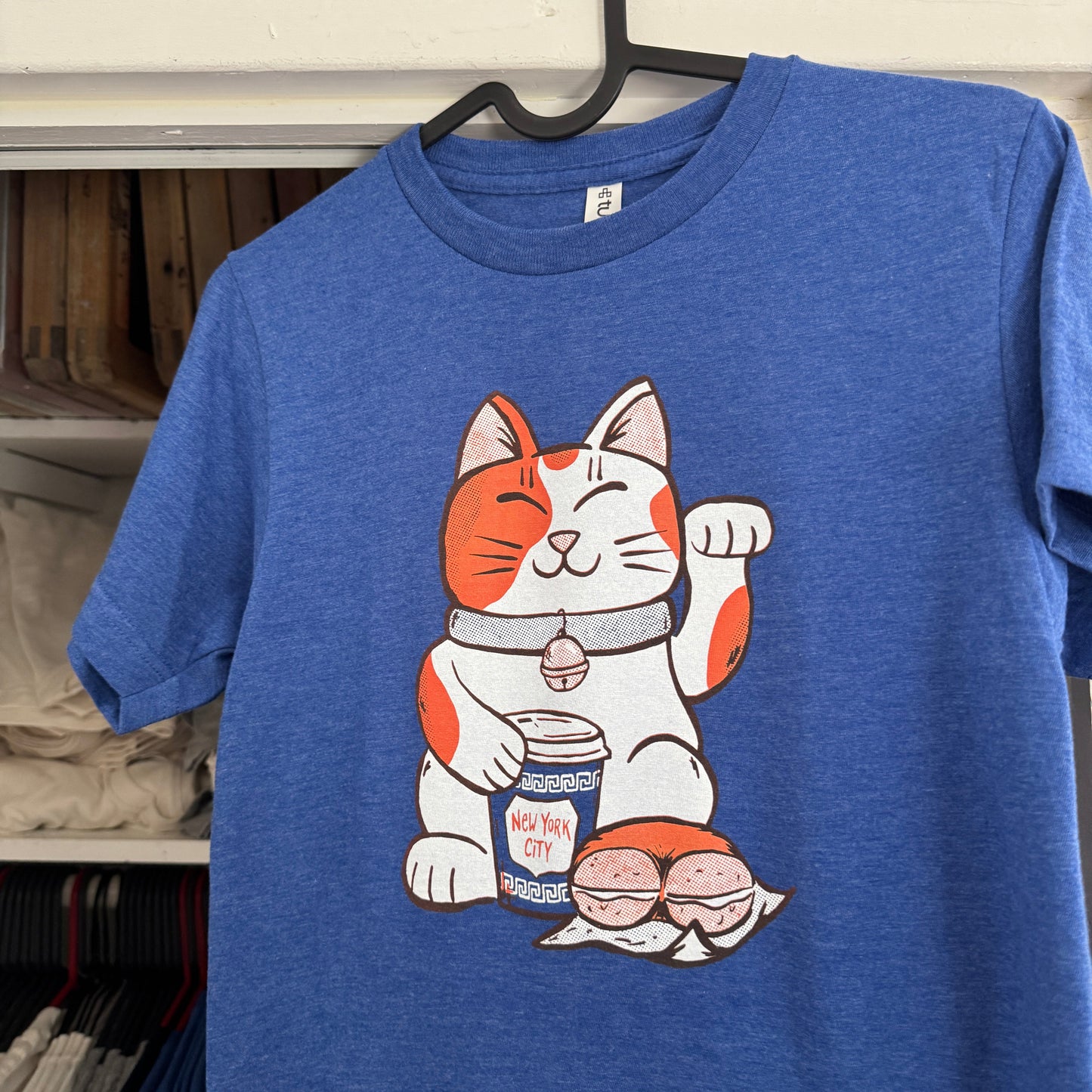 A blue heather t-shirt hanging on a black hanger, featuring a cartoon Maneki-neko cat graphic on the front with a NYC coffee cup and a bagel.