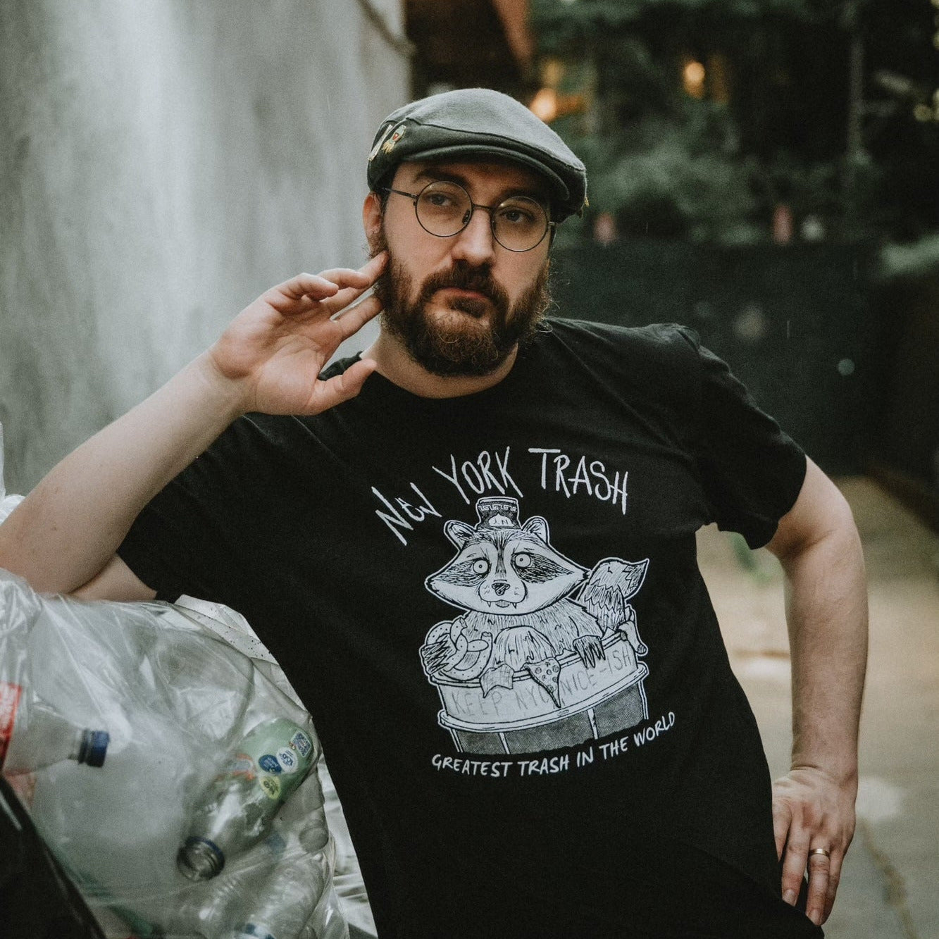 Man wearing a dark t-shirt with New York City Raccoon design and text, standing next to garbage.