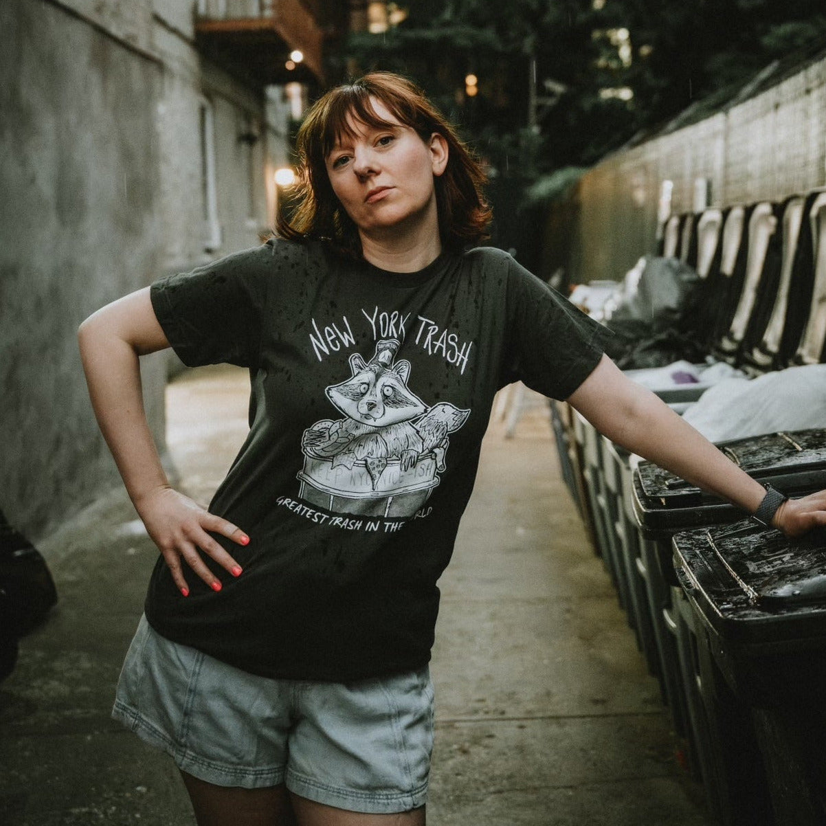 Man wearing a dark t-shirt with New York City Raccoon design and text, standing next to garbage.
